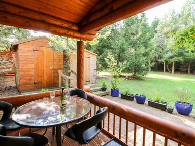 A covered wooden porch with a glass table and black chairs overlooking a garden with potted plants and two wooden sheds at Jalan End in Felton