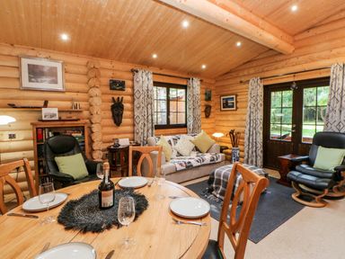 A dining and living area in a wooden log cabin with a round dining table set with plates and glasses two black armchairs a sofa with cushions a window and glass doors with curtains at Jalan End in Felton