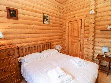 A bedroom with wooden log walls a wooden bed with white sheets and pillows a wooden dresser a wooden door and small tables with lamps at Jalan End in Felton