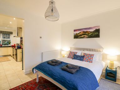 A bedroom with a double bed covered with a blue quilt and towels with an open doorway to a kitchen at The Hideaway in Malvern
