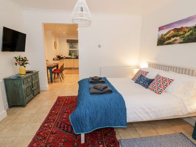 A bedroom with a bed, blue quilt and pillows, a green cabinet with flowers, and a view into a kitchen at The Hideaway in Malvern