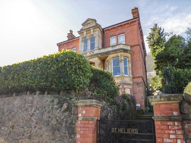 A red brick building with stone window frames behind a hedge and a stone wall with a gate reading St Heliers at The Hideaway in Malvern