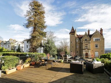 An outdoor wooden deck with a glass table and chairs and two cushioned armchairs with plants and buildings in the background at The Hideaway in Malvern