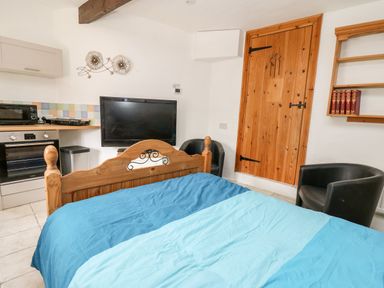 A bedroom with a double bed and blue bedding a kitchen area with an oven microwave and tiled backsplash a wooden door two black chairs a flat screen TV and a wall shelf with books at The Nook Star near Cenarth