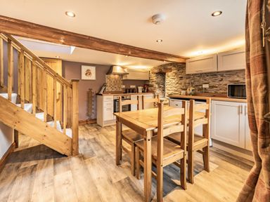 A kitchen with wooden dining table and chairs wooden stairs and white cabinets with stone wall at Pendre Cottage in Cenarth