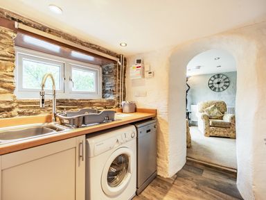 A kitchen with washing machine dishwasher and sink next to an archway leading to a sitting room with armchair and clock on the wall at Pendre Cottage in Cenarth