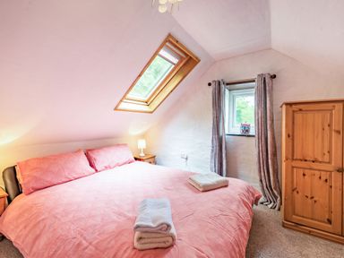 A bedroom with a double bed with pink bedding and folded towels on top a wooden wardrobe a small window with curtains and a skylight at Pendre Cottage in Cenarth