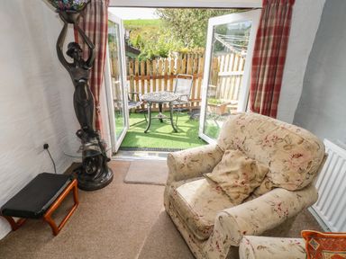 A sitting area with an armchair and a small table lamp next to open doors leading to a patio with a table and chairs at Pendre Cottage in Cenarth