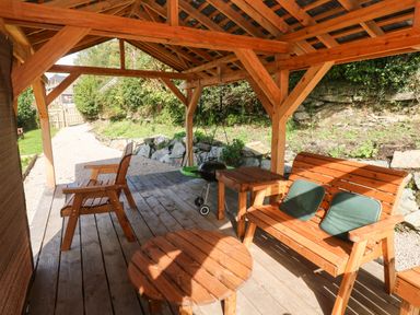 A wooden outdoor seating area with chairs table a small grill and greenery outside at Pendre Cottage in Cenarth
