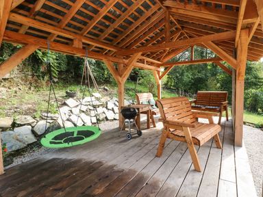An outdoor covered wooden patio with wooden benches a green swing and a charcoal grill at Pendre Cottage in Cenarth