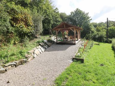 A wooden gazebo with outdoor furniture and a grill on a gravel path next to a grassy lawn surrounded by trees at Pendre Cottage in Cenarth