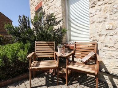 Two wooden chairs and a small table with a bottle, two glasses, and a packet against a stone wall outside at The Brewery in Brewers Quay Harbour