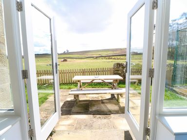 A wooden picnic table on a stone patio outside open glass doors with a picket fence and green field beyond at Greystead Institute in Bellingham
