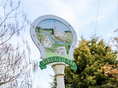 A white horseshoe-shaped sign with illustrations of buildings and a river with the word Greetham on a green banner at Rowan Lodge in Greetham Rutland
