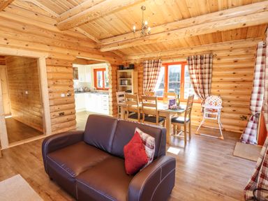 A dining area with wooden table and chairs next to windows with curtains and a leather sofa in a log cabin at Rowan Lodge in Greetham Rutland