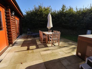 An outdoor patio with a wooden table and chairs set with an umbrella a lounge chair a wooden log cabin and a hot tub at Rowan Lodge in Greetham Rutland