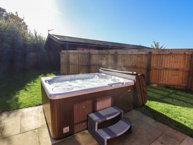An outdoor hot tub with steps on a patio next to a wooden fence with grass and a building in the background at Rowan Lodge in Greetham Rutland