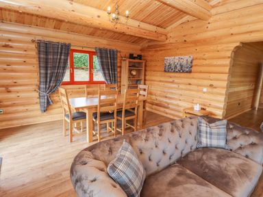 A wooden dining table with chairs near a window with curtains and a sofa with cushions in a log cabin living room at Maple Lodge in Greetham Rutland