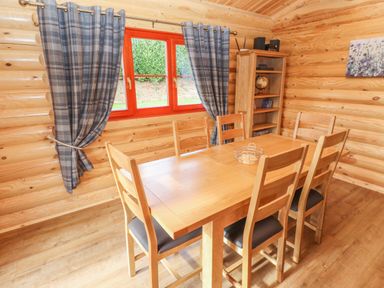 A dining room with a wooden table six chairs bookshelf and window with curtains at Maple Lodge in Greetham Rutland