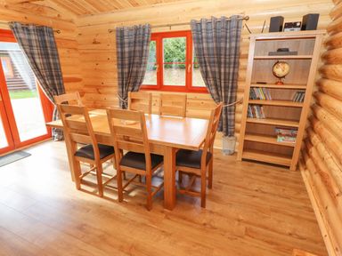 A dining area with a wooden table and six chairs next to windows with plaid curtains and a wooden bookshelf at Maple Lodge in Greetham Rutland