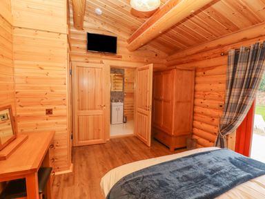 A bedroom with wooden walls ceiling and floor a bed with blue blanket a wooden wardrobe a dressing table and an open door showing a bathroom at Maple Lodge in Greetham Rutland
