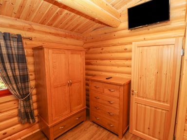 A wooden room with a wardrobe a chest of drawers a door a mounted television and a window with plaid curtains at Maple Lodge in Greetham Rutland
