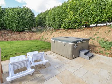 A patio with three white lounge chairs and a covered hot tub next to a grass lawn with hedges in the background at Maple Lodge in Greetham Rutland