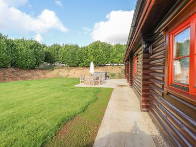 A garden with lawn and patio furniture next to a wooden cabin with red window frames at Maple Lodge in Greetham Rutland