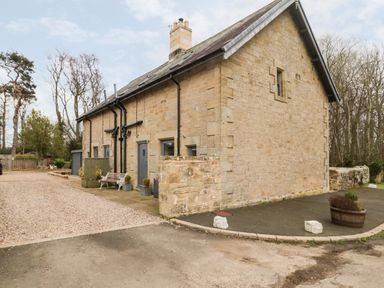 Exterior view of a stone building with multiple doors and benches near a gravel and paved driveway at Grange Cottages in Alnwick