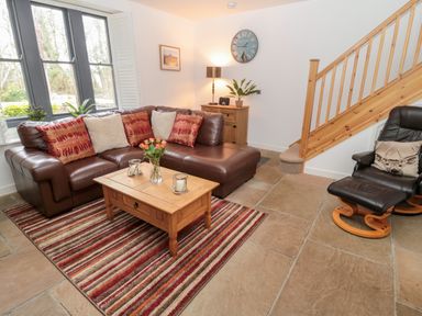A living room with a brown leather sectional sofa red and beige cushions a wooden coffee table striped rug a wooden sideboard with a lamp and clock and a leather armchair with a deer cushion next to wooden stairs at Grange Cottages in Alnwick