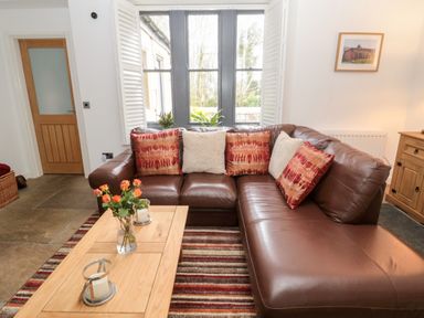 A brown leather sectional sofa with patterned and plain cushions near a wooden coffee table with flowers in a vase in a living room at Grange Cottages in Alnwick