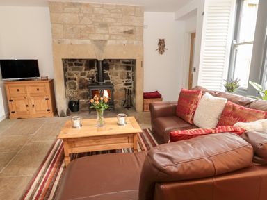 A living room with a stone fireplace a wood stove a brown leather sofa red cushions a wooden coffee table with flowers and candles a wooden cabinet with a television and a striped rug at Grange Cottages in Alnwick