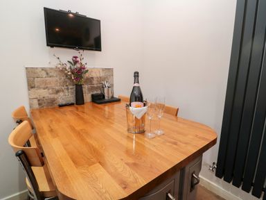 A dining area with a wooden table chairs a bottle of champagne in an ice bucket two champagne glasses a wall-mounted TV and flowers at Grange Cottages in Alnwick