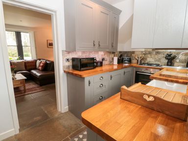 A kitchen with wooden countertops grey cabinets and kitchen appliances next to a doorway leading to a living room with a brown leather sofa at Grange Cottages in Alnwick