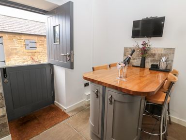 A kitchen area with a wooden countertop island, bar stools, a mounted TV, a half open stable door, and a bottle with glasses at Grange Cottages in Alnwick