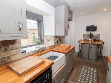 A kitchen with wooden countertops, a white sink, grey cabinets, a dining table with chairs, and a television on the wall at Grange Cottages in Alnwick