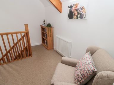 A small sitting area with a beige armchair and colorful pillow wooden bookshelf near wooden staircase and cow painting on wall at Grange Cottages in Alnwick