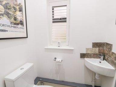 A small bathroom with a toilet a corner sink a window with blinds and a framed northumberland poster on the wall at Grange Cottages in Alnwick