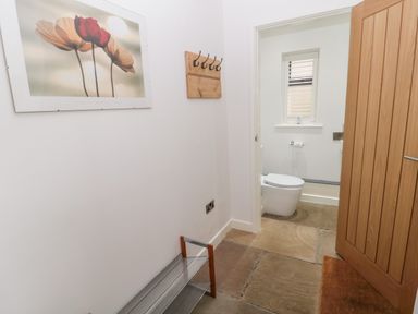 A hallway with a flower painting a wooden coat rack a metal shoe rack and an open door leading to a bathroom with a toilet and window at Grange Cottages in Alnwick