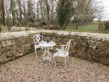 An outdoor patio area with two white metal chairs and a small round table with cups and a French press on a gravel floor surrounded by a stone wall at Grange Cottages in Alnwick