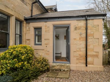 An exterior view of a stone building with a small entrance door and window surrounded by plants and gravel at Grange Cottages Alnwick