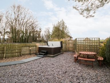 An outdoor area with a hot tub, wooden picnic table, wood chip ground, and fenced garden at Grange Cottages in Alnwick