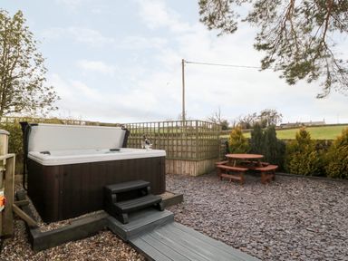 An outdoor area with a hot tub and a wooden picnic table surrounded by gravel and trees at Grange Cottages in Alnwick