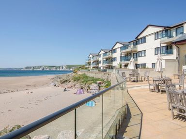 A beach with sand and people near a row of white buildings with balconies and patio tables at 17 Burgh Island Causeway in Bigbury-On-Sea