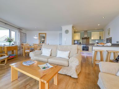 A living room with beige sofas a wooden coffee table dining table kitchen area and large window at 17 Burgh Island Causeway in Bigbury-On-Sea