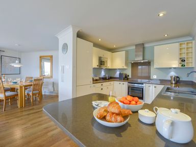 A kitchen with a countertop holding croissants, a bowl of fruit, and a teapot with a dining table in the background at 17 Burgh Island Causeway in Bigbury-On-Sea