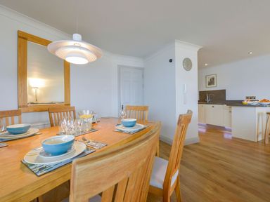 A dining area with wooden table and chairs set with bowls and glasses next to a kitchen with white cabinets at 17 Burgh Island Causeway in Bigbury-On-Sea