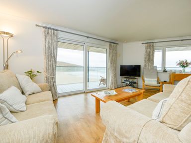 A living room with beige sofas a wooden coffee table a floor lamp a flat screen tv and large windows showing a beach view at 17 Burgh Island Causeway in Bigbury-On-Sea