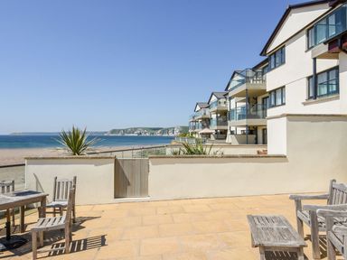A terrace with wooden chairs and tables overlooking the beach and sea with buildings along the coast at 17 Burgh Island Causeway in Bigbury-On-Sea