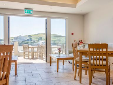 A dining area with wooden tables and chairs and a glass door leading to an outdoor seating area with a view of green hills at 17 Burgh Island Causeway in Bigbury-On-Sea
